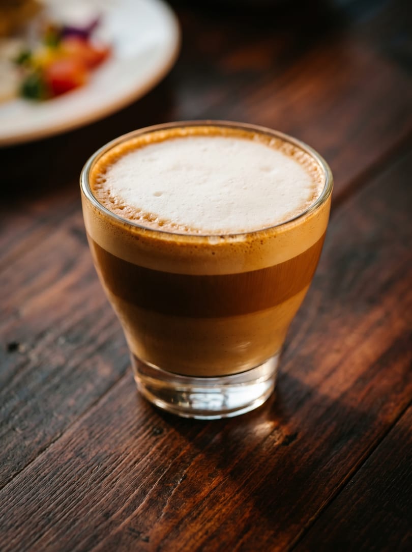 A glass of layered latte with frothy foam on a dark wooden table, with a blurred plate in the background.
